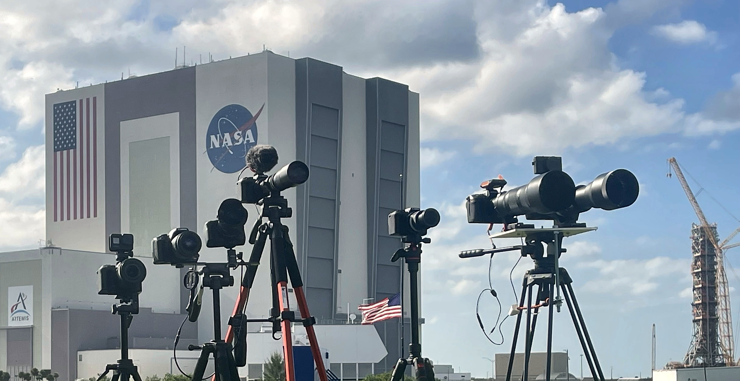 Robert Duvall's camera rigs on tripods, including cameras with long telephoto lenses, lined up in front of NASA's Vehicle Assembly Building, with a US flag at half-staff and a launch tower at right